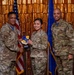 Three U.S. Air Force personnel in OCP (Operational Camouflage Pattern) uniforms pose for a formal award photo. The central figure is receiving a shield-shaped plaque bearing what appears to be the '7th Air Base Wing' emblem from the individual on the left, identified by nametape as 'HARRIS'. The ceremony takes place in front of an American flag and a blue Air Force flag against a gold tinsel backdrop.