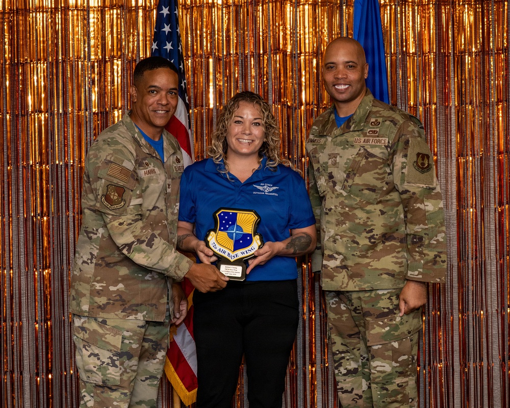Three individuals pose for a formal award photo against a gold tinsel backdrop. A civilian woman in a blue 'Air Force Outdoor Recreation' polo shirt holds a '72d Air Base Wing' shield-shaped plaque, flanked by two U.S. Air Force personnel in OCP (Operational Camouflage Pattern) uniforms. The setting appears to be a military awards ceremony.
