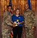Three individuals pose for a formal award photo against a gold tinsel backdrop. A civilian woman in a blue 'Air Force Outdoor Recreation' polo shirt holds a '72d Air Base Wing' shield-shaped plaque, flanked by two U.S. Air Force personnel in OCP (Operational Camouflage Pattern) uniforms. The setting appears to be a military awards ceremony.