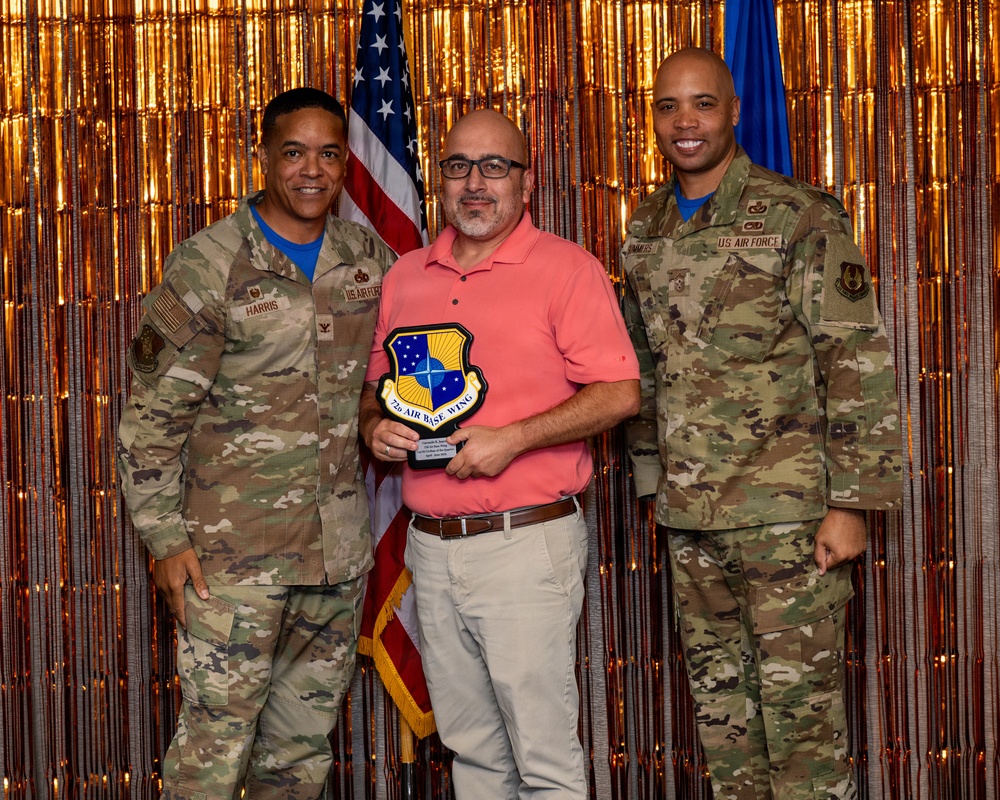 Three individuals pose for a formal award photo at what appears to be a U.S. Air Force recognition ceremony. The center person, dressed in civilian attire, holds a shield-shaped plaque bearing the '72d Air Base Wing' insignia. Two uniformed U.S. Air Force personnel in OCP (Operational Camouflage Pattern) uniforms flank him on either side.