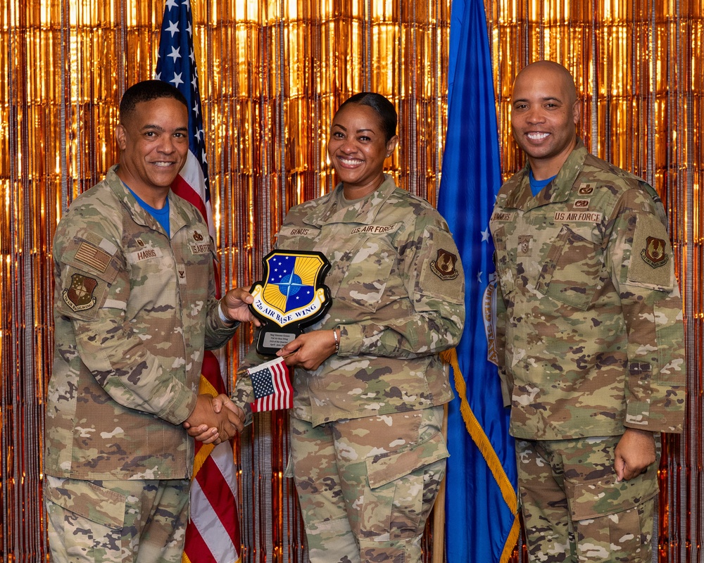 Three U.S. Air Force personnel in OCP (Operational Camouflage Pattern) uniforms pose for a formal recognition photo. The central figure is holding a shield-shaped plaque bearing the '7th Air Base Wing' insignia while shaking hands with the individual on the left. The ceremony appears to be an award or recognition event, set against a gold metallic backdrop with U.S. and Air Force flags.