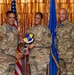 Three U.S. Air Force personnel in OCP (Operational Camouflage Pattern) uniforms pose for a formal recognition photo. The central figure is holding a shield-shaped plaque bearing the '7th Air Base Wing' insignia while shaking hands with the individual on the left. The ceremony appears to be an award or recognition event, set against a gold metallic backdrop with U.S. and Air Force flags.