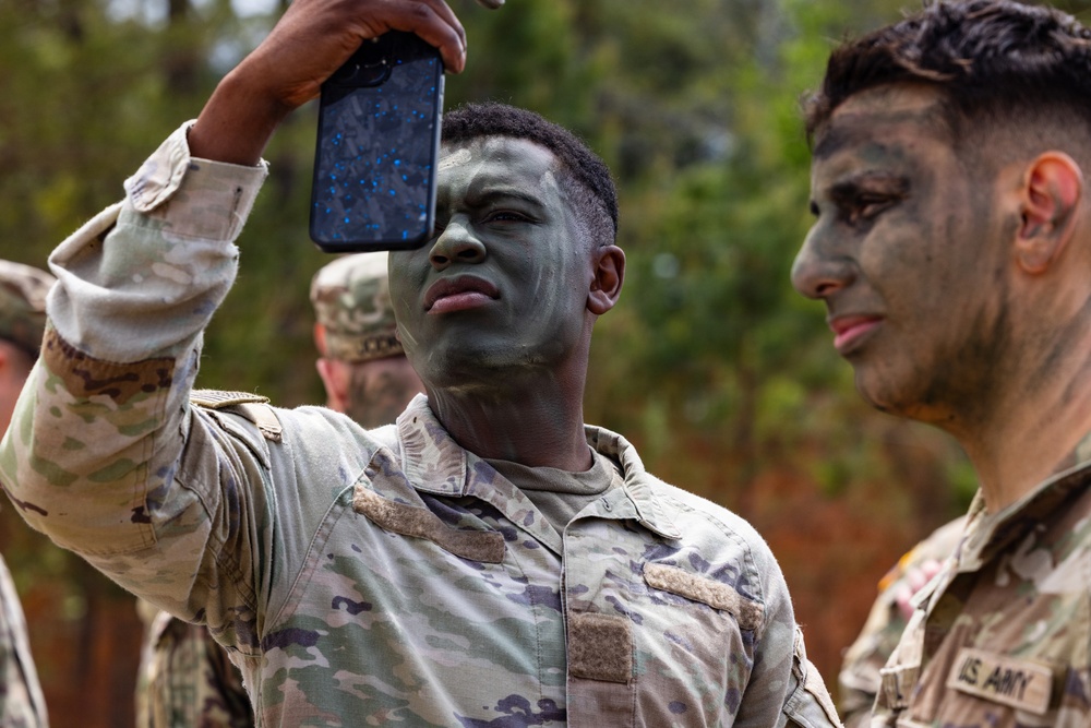 Two U.S. Army soldiers in MultiCam (OCP) uniforms are applying or checking camouflage face paint during what appears to be a field training exercise. The soldier in the foreground is using a smartphone as a mirror to inspect or apply green and black face paint. A second soldier with 'U.S. ARMY' tape visible on his uniform also has camouflage face paint applied.