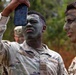 Two U.S. Army soldiers in MultiCam (OCP) uniforms are applying or checking camouflage face paint during what appears to be a field training exercise. The soldier in the foreground is using a smartphone as a mirror to inspect or apply green and black face paint. A second soldier with 'U.S. ARMY' tape visible on his uniform also has camouflage face paint applied.