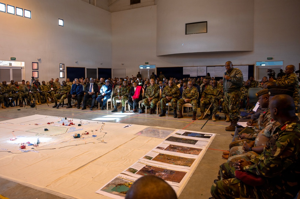 A large military briefing or operational planning session is taking place in a spacious indoor hall. A standing officer appears to be addressing a large audience of uniformed military personnel seated around a large floor map with tactical markers. Intelligence or reconnaissance documents and aerial imagery sheets are laid out on the floor alongside the map.