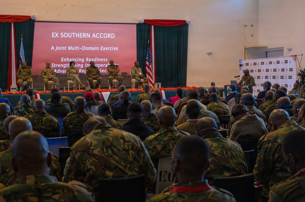 A formal military briefing or opening ceremony for 'Exercise Southern Accord,' described as a Joint Multi-Domain Exercise focused on enhancing readiness, strengthening interoperability, and advancing regional security. A large audience of military personnel in camouflage uniforms are seated facing a stage where senior officers are seated in a panel arrangement. A US flag and what appears to be an African nation's flag are visible on stage, indicating a multinational military exercise.