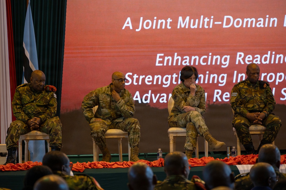 Four military personnel in camouflage uniforms are seated on a stage panel at a formal military conference or symposium. The presentation screen behind them partially displays text referencing 'A Joint Multi-Domain' operation and themes of 'Enhancing Readiness,' 'Strengthening Interoperability,' and 'Advancing Regional Security.' An audience of uniformed personnel is visible in the foreground.
