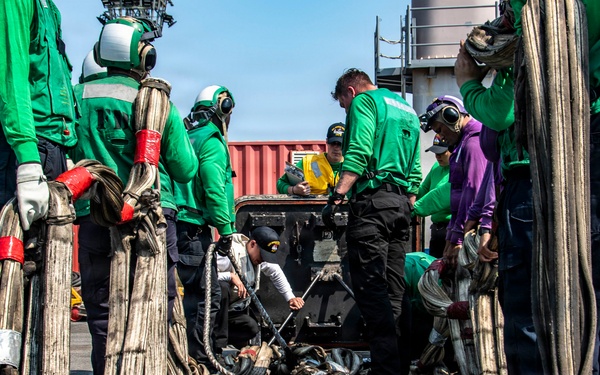 A group of workers, predominantly wearing green shirts and safety gear, are handling large bundles of heavy cables or hoses on what appears to be a naval vessel or industrial dock. The cables are bundled and marked with red and silver tape at intervals, suggesting organized rigging or mooring operations. One individual in a purple shirt stands out among the green-clad crew, possibly indicating a supervisory or specialist role.
