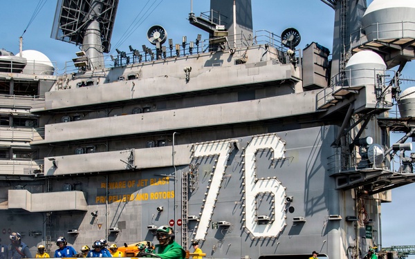 The image shows the flight deck of USS Ronald Reagan (CVN-76), a Nimitz-class nuclear-powered aircraft carrier, with crew members conducting flight deck operations. A white aircraft tow tractor is being operated near the island superstructure, which prominently displays the hull number '76'. Personnel in color-coded jerseys indicating their roles are visible across the deck.
