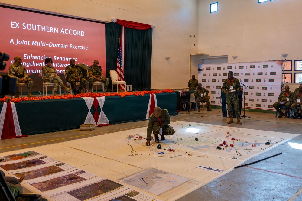 A military briefing or exercise demonstration for 'Ex Southern Accord - A Joint Multi-Domain Exercise' is taking place in a large indoor hall. Personnel in military uniforms are gathered around a large floor map with tactical markers, while a speaker addresses the audience from a podium. A dignitary panel is seated at a decorated head table beneath a presentation screen and an American flag.