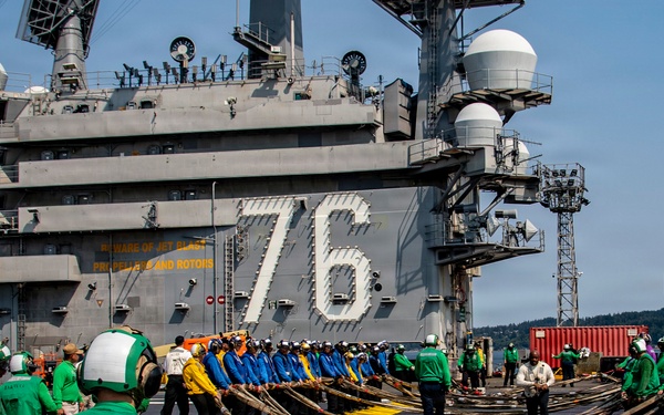 Personnel are conducting flight deck operations aboard USS Ronald Reagan (CVN-76), a Nimitz-class aircraft carrier. Crew members in color-coded jerseys are handling what appear to be arresting wire cables or similar deck equipment in a coordinated formation. The ship is docked at a naval installation, identifiable by the hull number '76' prominently displayed on the island superstructure.