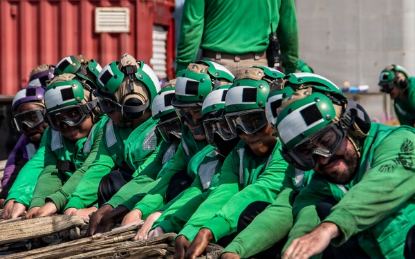 A group of naval personnel dressed in green flight deck jerseys and helmets with goggles are crouched low on what appears to be an aircraft carrier flight deck, collectively handling large bundled cables or arresting gear wires. The crew members are working in a coordinated team effort, pulling or managing heavy equipment on the deck surface. In the background, additional personnel in yellow and green jerseys are visible near red shipping containers or deck structures.