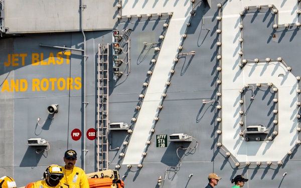 A large group of naval personnel are conducting what appears to be a fire hose drill or equipment handling exercise on the flight deck of USS Ronald Reagan (CVN-76), a Nimitz-class aircraft carrier. Crew members wearing color-coded jerseys (yellow, blue, green) and protective helmets with visors are crouched in a line managing large fire hoses or arresting gear cables. The ship's hull number '76' and safety warning text are prominently visible on the island superstructure.