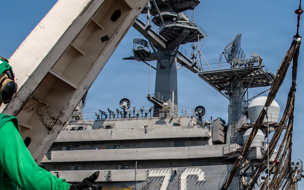 The image shows the flight deck of USS Ronald Reagan (CVN-76), a Nimitz-class aircraft carrier, with deck crew in green jerseys conducting flight deck operations. A large arresting wire or net assembly is visible being handled on the deck, suspended by a crane or davit arm. The ship's island superstructure with hull number '76' is prominently visible in the background.