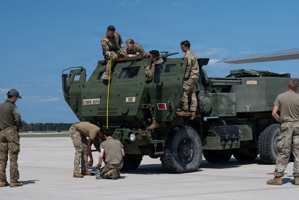 Military personnel in camouflage uniforms are conducting measurements and inspections on a HIMARS (High Mobility Artillery Rocket System) vehicle marked '272BSG 182FA' and 'B21' on an airfield tarmac. The activity appears to involve aircraft load planning or pre-airlift preparation, with personnel using a measuring tape on top of and around the vehicle. An aircraft wing tip is visible in the upper right corner, suggesting the vehicle is being prepared for air transport.
