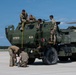 Military personnel in camouflage uniforms are conducting measurements and inspections on a HIMARS (High Mobility Artillery Rocket System) vehicle marked '272BSG 182FA' and 'B21' on an airfield tarmac. The activity appears to involve aircraft load planning or pre-airlift preparation, with personnel using a measuring tape on top of and around the vehicle. An aircraft wing tip is visible in the upper right corner, suggesting the vehicle is being prepared for air transport.