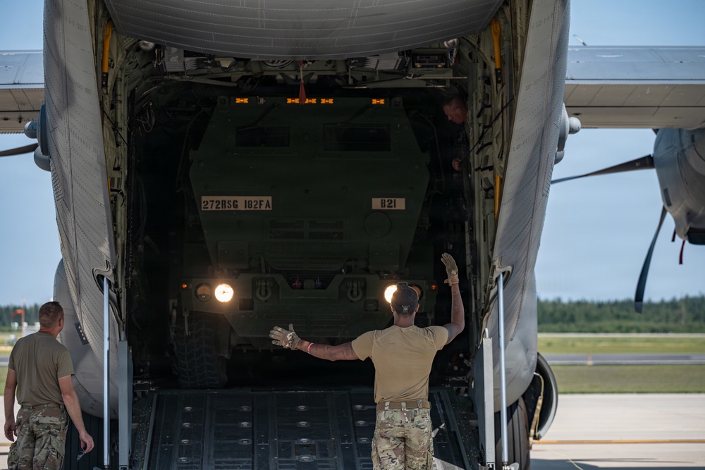 Military personnel are guiding a tactical wheeled vehicle out of a large military transport aircraft via its rear cargo ramp. The vehicle, marked '272RSG 182FA' and 'B21', appears to be a military truck being offloaded onto an airfield tarmac. At least two ground crew members are visible directing the operation using hand signals.