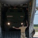 Military personnel are guiding a tactical wheeled vehicle out of a large military transport aircraft via its rear cargo ramp. The vehicle, marked '272RSG 182FA' and 'B21', appears to be a military truck being offloaded onto an airfield tarmac. At least two ground crew members are visible directing the operation using hand signals.