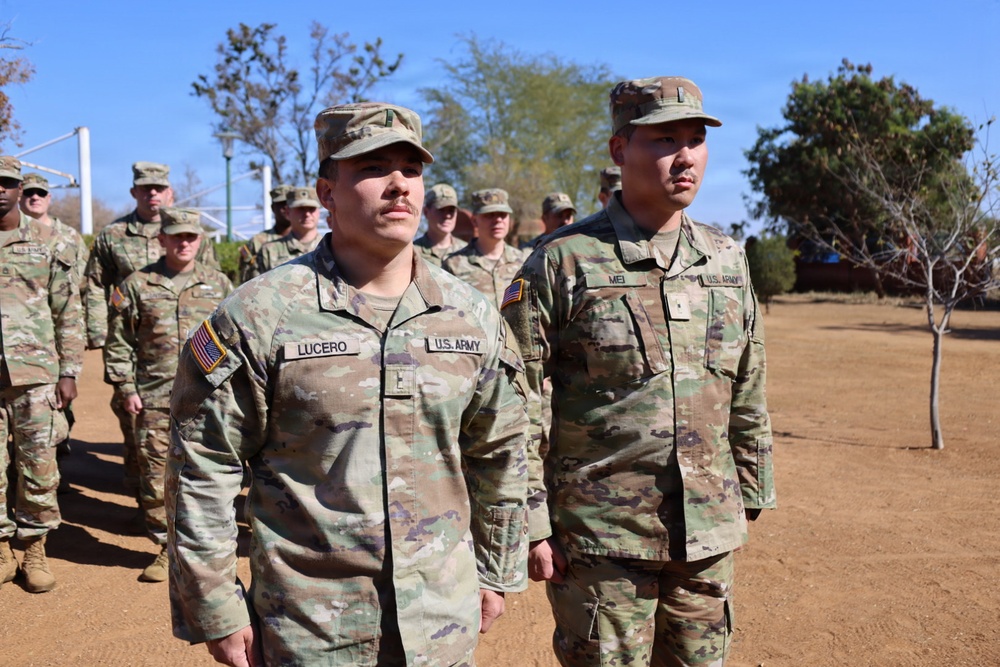 A group of U.S. Army soldiers in Operational Camouflage Pattern (OCP) uniforms are standing in formation outdoors on a dirt surface. Two soldiers are prominently visible in the foreground, identified by name tapes as 'LUCERO' and 'MEI', both wearing U.S. Army insignia and American flag patches. The setting appears to be a military installation or training area with trees and blue sky in the background.