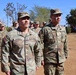 A group of U.S. Army soldiers in Operational Camouflage Pattern (OCP) uniforms are standing in formation outdoors on a dirt surface. Two soldiers are prominently visible in the foreground, identified by name tapes as 'LUCERO' and 'MEI', both wearing U.S. Army insignia and American flag patches. The setting appears to be a military installation or training area with trees and blue sky in the background.