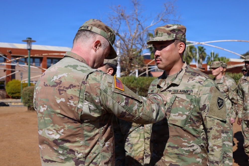 A U.S. Army ceremony is taking place outdoors, where a soldier with the name tape 'АМПION' (likely 'CHAMPION') is pinning a medal or insignia onto a soldier identified by the name tape 'ME' (partially visible) and 'U.S. ARMY' tape. Both soldiers are wearing Operational Camouflage Pattern (OCP) uniforms. The setting appears to be a military installation, possibly in a warm-climate region based on the palm trees and vegetation.