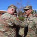 A U.S. Army ceremony is taking place outdoors, where a soldier with the name tape 'АМПION' (likely 'CHAMPION') is pinning a medal or insignia onto a soldier identified by the name tape 'ME' (partially visible) and 'U.S. ARMY' tape. Both soldiers are wearing Operational Camouflage Pattern (OCP) uniforms. The setting appears to be a military installation, possibly in a warm-climate region based on the palm trees and vegetation.