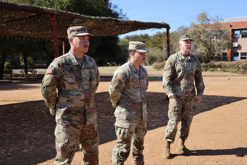 Three U.S. Army soldiers in Operational Camouflage Pattern (OCP) uniforms are standing and walking on a dirt surface at what appears to be a military installation or training area. The soldiers appear to be engaged in conversation or a briefing outdoors. Name tapes are partially visible on at least two of the soldiers, with 'MEI' and 'LUCERO' legible.