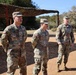Three U.S. Army soldiers in Operational Camouflage Pattern (OCP) uniforms are standing and walking on a dirt surface at what appears to be a military installation or training area. The soldiers appear to be engaged in conversation or a briefing outdoors. Name tapes are partially visible on at least two of the soldiers, with 'MEI' and 'LUCERO' legible.