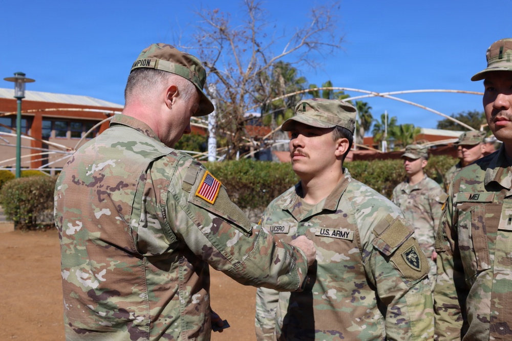 A U.S. Army ceremony is taking place outdoors, where a soldier with the name tape 'CHAMPION' is pinning or presenting something to a soldier named 'LUCERO'. Both wear Operational Camouflage Pattern (OCP) uniforms with U.S. Army insignia. Additional soldiers are visible in formation in the background.