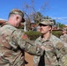 A U.S. Army ceremony is taking place outdoors, where a soldier with the name tape 'CHAMPION' is pinning or presenting something to a soldier named 'LUCERO'. Both wear Operational Camouflage Pattern (OCP) uniforms with U.S. Army insignia. Additional soldiers are visible in formation in the background.
