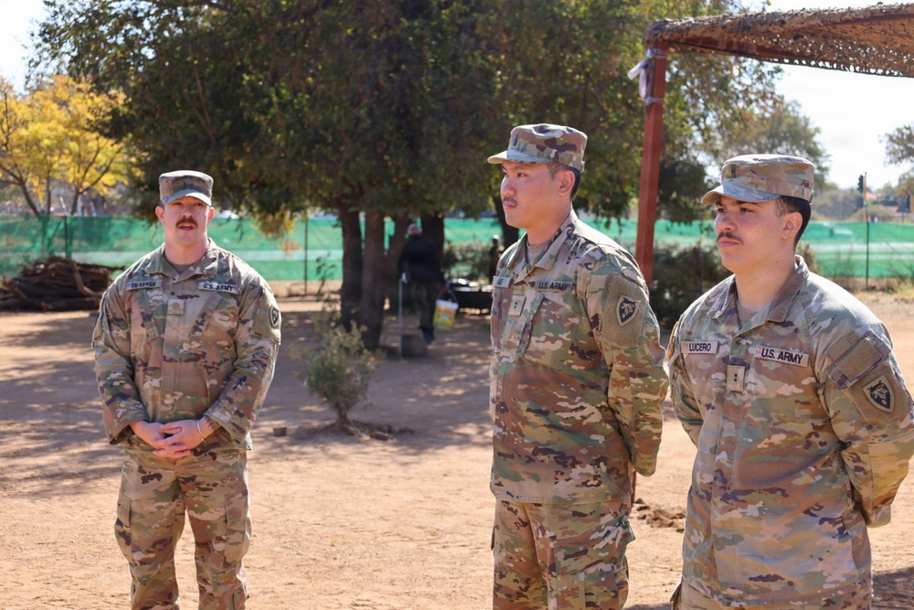 Three U.S. Army soldiers in OCP (Operational Camouflage Pattern) uniforms are standing outdoors in what appears to be a field or training environment. The soldiers are at attention or in a relaxed standing position, possibly receiving a briefing or participating in a formation. Name tapes visible include 'LUCERO' on the right soldier and a partially visible name on the left soldier.