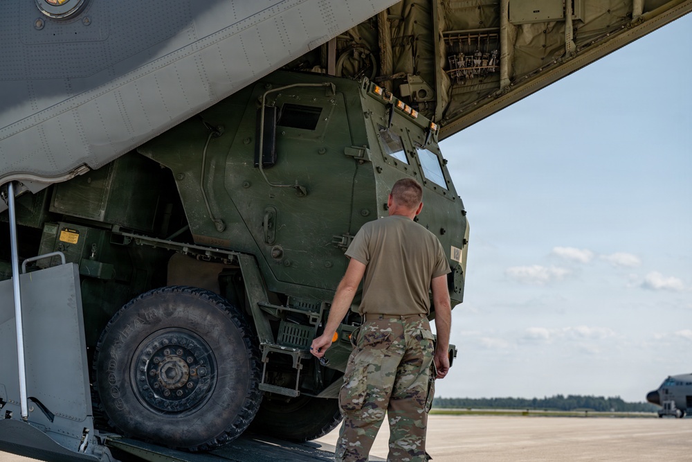 A military service member in OCP (Operational Camouflage Pattern) uniform is inspecting or overseeing the loading/unloading of a large military tactical vehicle into or out of a military transport aircraft. The vehicle appears to be an M142 HIMARS (High Mobility Artillery Rocket System) or similar heavy military truck being transported via what appears to be a C-17 Globemaster III or similar large cargo aircraft. The scene takes place on a military airfield tarmac under clear skies.