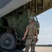 A military service member in OCP (Operational Camouflage Pattern) uniform is inspecting or overseeing the loading/unloading of a large military tactical vehicle into or out of a military transport aircraft. The vehicle appears to be an M142 HIMARS (High Mobility Artillery Rocket System) or similar heavy military truck being transported via what appears to be a C-17 Globemaster III or similar large cargo aircraft. The scene takes place on a military airfield tarmac under clear skies.