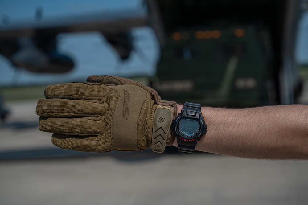A close-up photograph of a person's forearm wearing a tan/coyote brown tactical glove on one hand and a Casio G-Shock digital watch on the wrist. The background shows what appears to be a military airfield environment with a large aircraft and military vehicle visible in soft focus. The image appears to be taken in an operational military or aviation context.