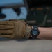 A close-up photograph of a person's forearm wearing a tan/coyote brown tactical glove on one hand and a Casio G-Shock digital watch on the wrist. The background shows what appears to be a military airfield environment with a large aircraft and military vehicle visible in soft focus. The image appears to be taken in an operational military or aviation context.