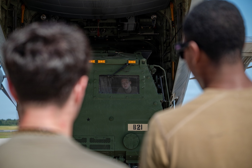 Two military personnel in tan/olive uniforms are observing a green military vehicle marked 'B21' being loaded or unloaded from the rear cargo ramp of a large military transport aircraft. A third person is visible through the windshield of the vehicle, operating it. The scene appears to take place on a military airfield.