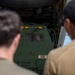 Two military personnel in tan/olive uniforms are observing a green military vehicle marked 'B21' being loaded or unloaded from the rear cargo ramp of a large military transport aircraft. A third person is visible through the windshield of the vehicle, operating it. The scene appears to take place on a military airfield.