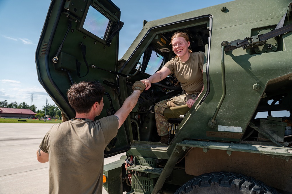 Two military personnel in olive drab uniforms are interacting beside a large military tactical vehicle, likely an LMTV or similar Army truck. A female soldier seated in the cab of the vehicle is smiling and shaking hands or fist-bumping with a male soldier standing on the ground. The setting appears to be a military installation or airfield on a clear, sunny day.