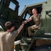 Two military personnel in olive drab uniforms are interacting beside a large military tactical vehicle, likely an LMTV or similar Army truck. A female soldier seated in the cab of the vehicle is smiling and shaking hands or fist-bumping with a male soldier standing on the ground. The setting appears to be a military installation or airfield on a clear, sunny day.