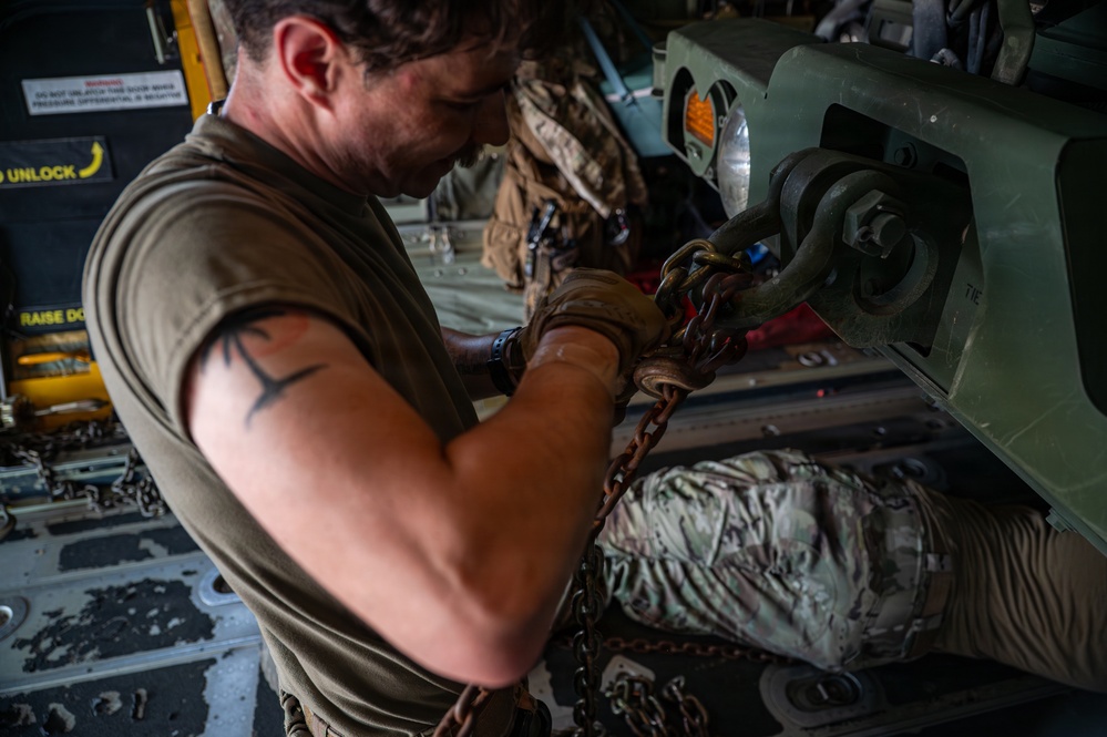 A military service member in a olive drab sleeveless shirt is securing cargo chains to a tie-down point inside what appears to be a military transport aircraft, likely a C-130 Hercules. The individual has a barbed wire tattoo on their upper arm and is wearing tactical gloves while working with heavy chain rigging. Military gear and equipment bags in MultiCam/OCP camouflage pattern are visible nearby.