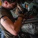 A military service member in a olive drab sleeveless shirt is securing cargo chains to a tie-down point inside what appears to be a military transport aircraft, likely a C-130 Hercules. The individual has a barbed wire tattoo on their upper arm and is wearing tactical gloves while working with heavy chain rigging. Military gear and equipment bags in MultiCam/OCP camouflage pattern are visible nearby.