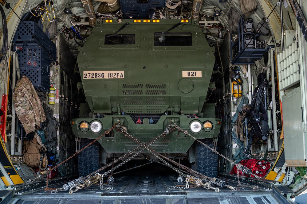 A military wheeled vehicle marked '272RSG 182FA' and 'B21' is secured inside the cargo hold of a large military transport aircraft, likely a C-130 Hercules. The vehicle, which appears to be an M1083 FMTV or similar tactical truck variant, is restrained with heavy chains in an X-pattern across the cargo floor. The interior of the aircraft shows typical loadmaster equipment, tie-down rails, and various gear stowed along the fuselage walls.