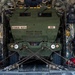 A military wheeled vehicle marked '272RSG 182FA' and 'B21' is secured inside the cargo hold of a large military transport aircraft, likely a C-130 Hercules. The vehicle, which appears to be an M1083 FMTV or similar tactical truck variant, is restrained with heavy chains in an X-pattern across the cargo floor. The interior of the aircraft shows typical loadmaster equipment, tie-down rails, and various gear stowed along the fuselage walls.
