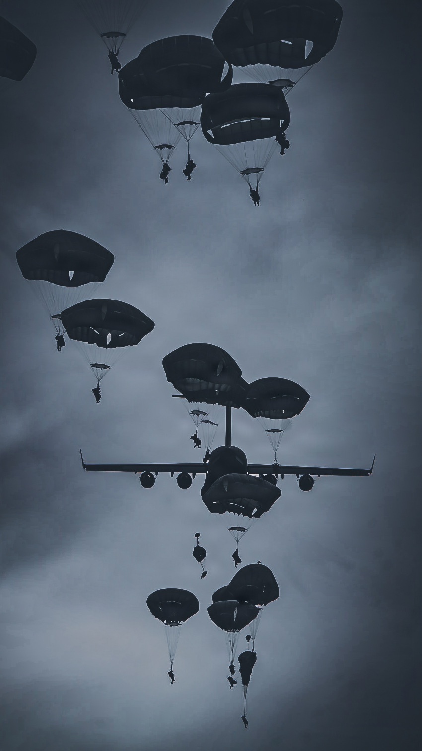 A low-angle upward shot captures a large military transport aircraft dispensing numerous paratroopers against an overcast sky. Multiple parachutes at various stages of deployment are visible both above and below the aircraft, indicating an active mass tactical airborne operation. The monochromatic, dark tonal quality of the image gives it a dramatic, silhouetted appearance.