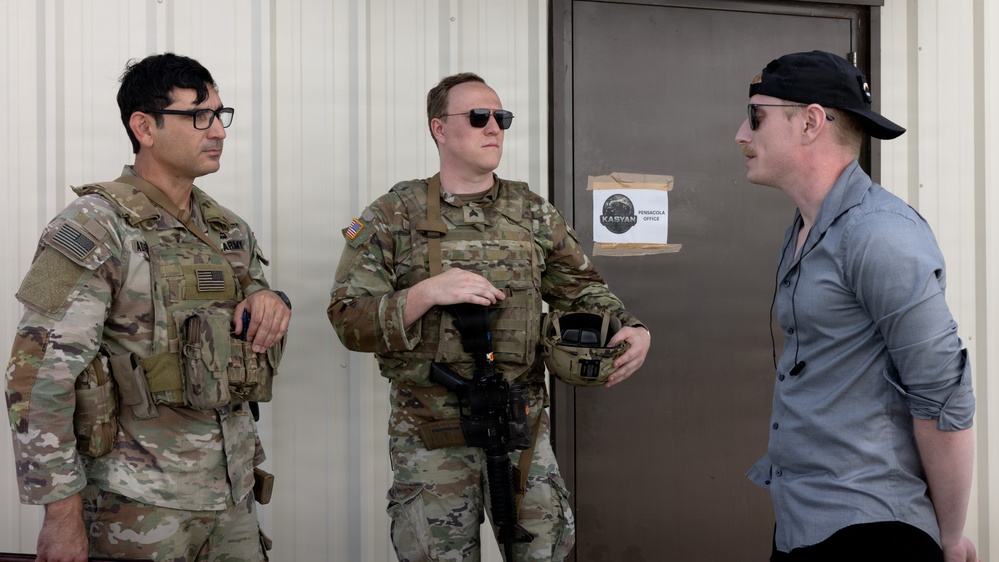 Two U.S. Army soldiers in MultiCam (OCP) uniforms with body armor and tactical gear are conversing with a civilian man wearing a gray shirt and black baseball cap. The interaction takes place outside a building near a door with a sign reading 'KASYAN PENSACOLA OFFICE'. The setting appears to be a military installation or training facility.