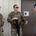 Two U.S. Army soldiers in MultiCam (OCP) uniforms with body armor and tactical gear are conversing with a civilian man wearing a gray shirt and black baseball cap. The interaction takes place outside a building near a door with a sign reading 'KASYAN PENSACOLA OFFICE'. The setting appears to be a military installation or training facility.