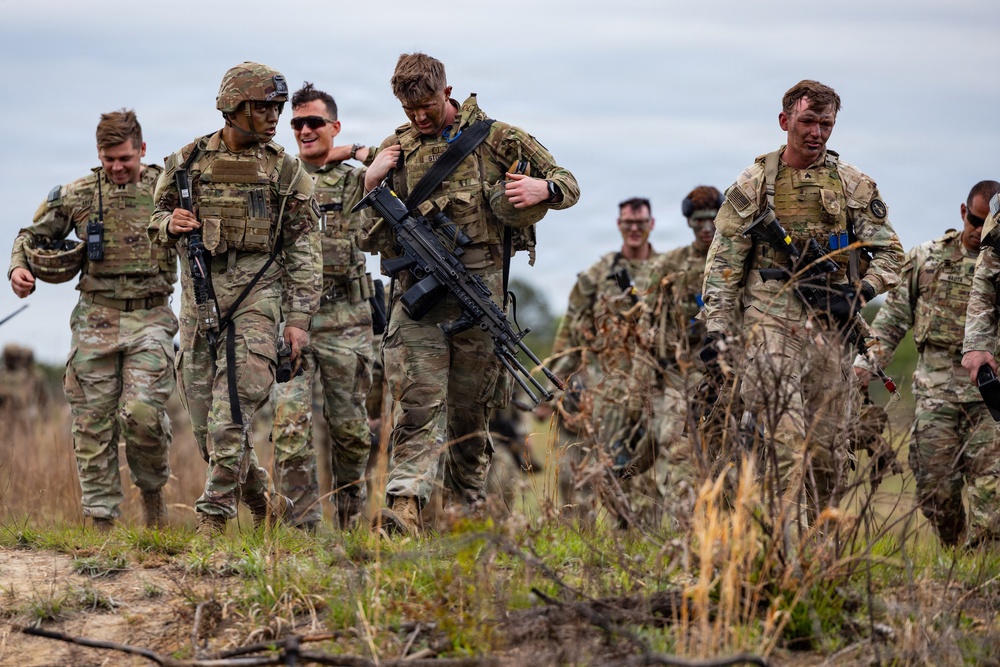 A group of military personnel in full combat gear are walking across an open field, appearing to be during or after a training exercise. The soldiers are wearing MultiCam/OCP camouflage uniforms and carrying various weapons and tactical equipment. Several soldiers appear fatigued but in good spirits, with some smiling and supporting one another.