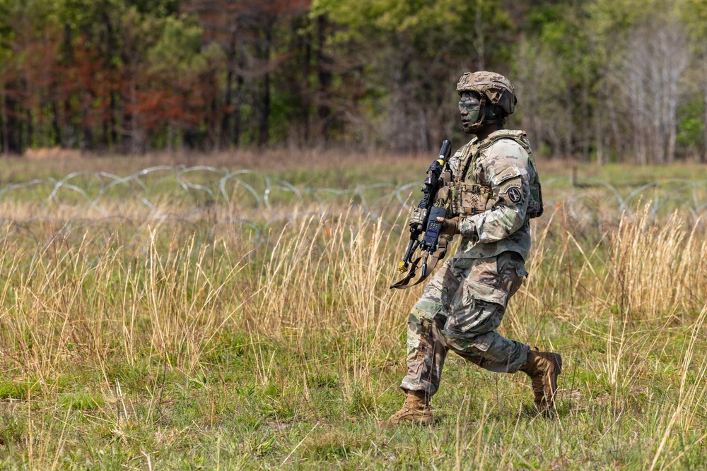 A U.S. Army soldier in full combat gear is moving across an open grassy field during what appears to be a military training exercise. The soldier is wearing OCP (Operational Camouflage Pattern) uniform with face paint applied, carrying a rifle, and moving past a concertina wire obstacle. The setting appears to be a military training area with mixed terrain including tall dry grass and a tree line in the background.