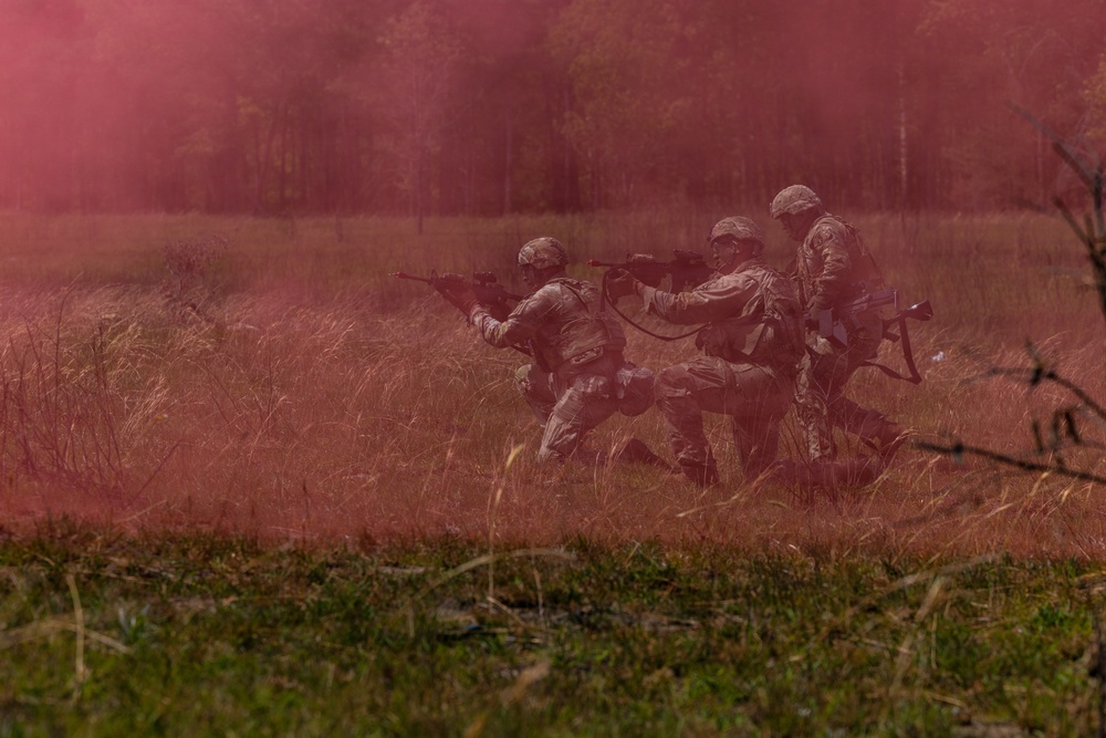 Three soldiers in camouflage uniforms and helmets are advancing through a field obscured by red/pink smoke, likely during a military training exercise. They are moving in a tactical formation while carrying rifles in a ready position. The scene takes place in an open grassy field with a tree line visible in the background.