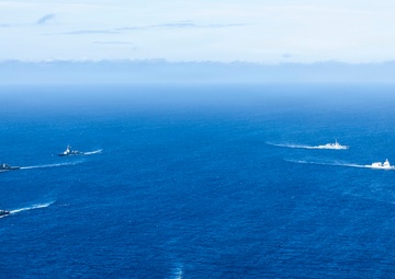 An aerial photograph shows a formation of seven naval warships traversing open blue ocean waters. The vessels are arranged in a deliberate tactical formation, suggesting a coordinated multinational or fleet exercise. All ships are underway and producing visible wake trails.